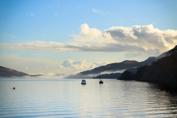 Looking South on the West shore of Loch Lomond, little boats float in front of cloud-covered hills.